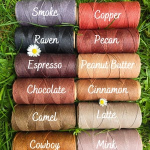 May include: Ten cylindrical spools of thread arranged in two columns, labeled with the colors Smoke, Copper, Raven, Pecan, Espresso, Peanut Butter, Chocolate, Cinnamon, Camel, Latte, Cowboy, and Mink, featuring a white daisy flower.