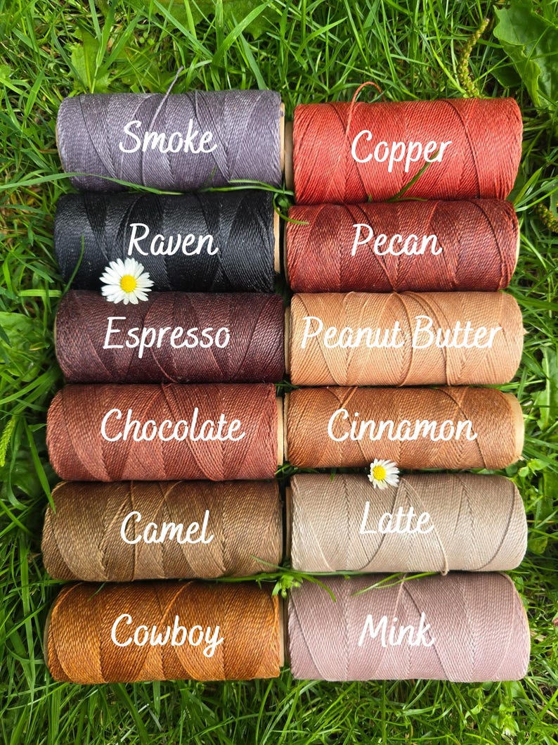 May include: Ten cylindrical spools of thread arranged in two columns, labeled with the colors Smoke, Copper, Raven, Pecan, Espresso, Peanut Butter, Chocolate, Cinnamon, Camel, Latte, Cowboy, and Mink, featuring a white daisy flower.