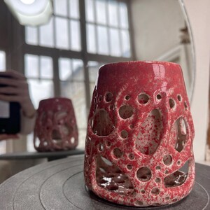 Puede incluir: Un jarrón de cerámica hecho a mano con un esmalte moteado rojo. El jarrón tiene forma cilíndrica con numerosas aberturas circulares, que revelan el interior. El jarrón se exhibe sobre una rueda de alfarero gris, con un reflejo en un espejo cercano.
