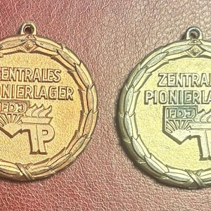 May include: Two circular medals with the text "ZENTRALES PIONIERLAGER" and an emblem. One medal is gold-toned, the other is silver-toned. Both have a wreath border and a loop for hanging.