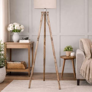 May include: A wooden tripod floor lamp with a beige fabric shade. The lamp has a natural wood finish and stands on a light-colored rug. The room also features a side table, sofa, and a vase of white flowers.