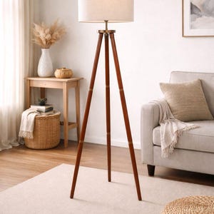 May include: A wooden tripod floor lamp with a beige drum shade. The lamp has a dark brown finish on the legs and brass-colored accents. The lamp is in a living room setting with a neutral color palette.