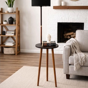 May include: A modern floor lamp with a black lampshade and a dark brown wooden pole. It has a small, round side table with three wooden legs. A small Buddha statue and a book are on the table. The lamp is in a living room setting.