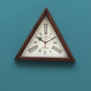 May include: A triangular wall clock with a dark brown wooden frame and an off-white face. The clock features black Roman numerals, black ornate hands, a red second hand, and the text Jones & London EST 1923 and OXFORD printed on the face.