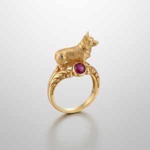 May include: A gold-colored ring featuring a detailed corgi dog figurine atop a decorative band. The ring has a round, red gemstone at the center of the band. The ring is a unique piece of jewelry.