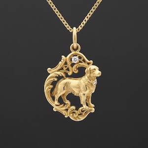 Op de afbeelding: Een goudkleurige hangerketting met een gedetailleerd hondenontwerp in een sierlijk frame. De hanger heeft een kleine diamantaccent op de halsband van de hond en een andere aan de bovenkant van het frame. De ketting is ook goudkleurig.