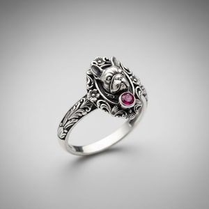 May include: A silver ring featuring a detailed French Bulldog design within an oval frame, accented with floral motifs. The ring includes a small, round, red gemstone. The band has an engraved pattern.