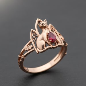 May include: Rose gold ring featuring a cat design with bat-wing accents. The cat sits atop a teardrop-shaped, deep red gemstone. The ring has intricate filigree details and a smooth, polished finish.