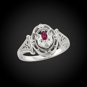 May include: A silver ring featuring a cat face design with a ruby-colored gemstone. The ring has an ornate, vintage-inspired setting with detailed filigree and geometric patterns on the band. The cat's face is centered within an oval frame.