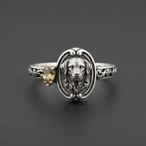 May include: A silver ring featuring a detailed Labrador Retriever head within an oval frame. The ring has a teardrop-shaped, amber-colored gemstone on the side. The band is decorated with floral patterns.