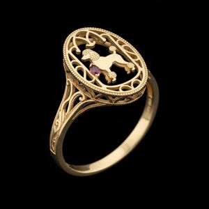 May include: A gold ring featuring an oval design with a poodle silhouette and a small red gemstone. The ring has an ornate, filigree-style setting and a polished band, set against a black background.