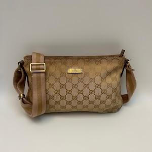 Puede incluir: Un bolso de hombro de lona en color beige y marrón con un patrón de monograma GG repetitivo. El bolso incluye una correa de tela ancha y ajustable a rayas en tonos rosa y beige, herrajes de metal dorado y una placa rectangular dorada con el logotipo de Gucci.