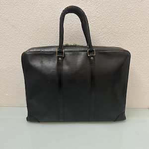 May include: A black briefcase with a textured horizontal line pattern, featuring two vertical leather straps, two top handles, and a zippered closure with metallic hardware. The rectangular bag has reinforced corners and a structured design.