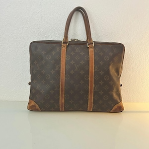 May include: Brown monogrammed briefcase with leather accents. The bag features a top handle, two vertical leather straps, and leather corner protectors. The bag has gold-toned hardware and a zipper closure. The bag is rectangular and has a classic design.