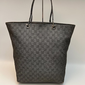 Puede incluir: Un bolso tote de color gris oscuro con un patrón de monograma GG negro repetido en el exterior. El bolso incluye dos asas finas de color gris oscuro sujetas con herrajes metálicos cuadrados de tono plateado en el borde superior.