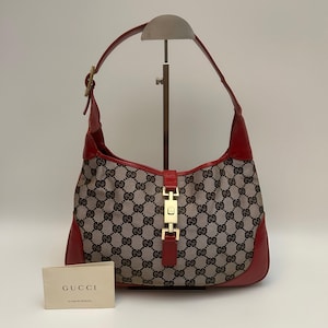 Puede incluir: Un bolso hobo de Gucci con cuerpo de lona beige y patrón de monograma GG en marrón oscuro, acentuado con ribetes de cuero rojo y un cierre de pistón de metal dorado. Una pequeña tarjeta blanca con el texto GUCCI se sitúa frente al bolso.