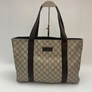Puede incluir: Un bolso tote de color beige y marrón con un patrón de monograma GG repetido en el exterior. El bolso incluye dos correas verticales de tela marrón oscuro que se extienden hasta las asas y un pequeño parche rectangular de cuero marrón.