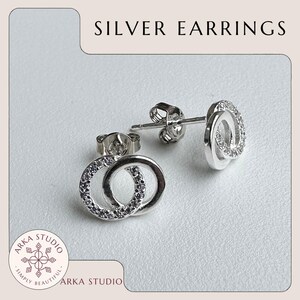 May include: Silver earrings with a double-circle design, each circle encrusted with small, clear stones. The earrings are displayed on a light background. The text "SILVER EARRINGS" is at the top of the image, and the "ARKA STUDIO" logo is at the bottom.
