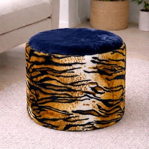 Könnte beinhalten: Ein zylindrischer Hocker mit einer plüschigen, dunkelblauen Oberfläche und Seiten, die mit weichem Kunstfell bezogen sind, das ein realistisches Tigerstreifenmuster in Orange-, Schwarz- und Weißtönen aufweist und eine strukturierte Optik erzeugt.