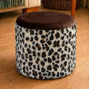Puede incluir: Un taburete cilíndrico con una parte superior circular de felpa marrón oscuro y laterales cubiertos de tela de piel sintética con un estampado de leopardo en negro y marrón. El taburete tiene una base plana y una forma redonda uniforme.