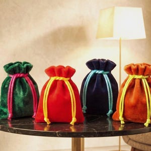 Op de afbeelding: Vier fluwelen trekkoordzakken in groen, rood, marineblauw en oranje. Elke tas heeft een contrasterende koordsluiting in roze, geel, lichtblauw en geel. De tassen worden gepresenteerd op een zwarte marmeren tafel.