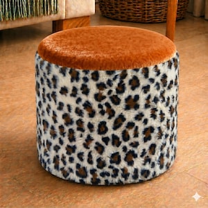 Könnte beinhalten: Ein runder Hocker mit einem weichen, orangebraunen Oberteil und einer zylindrischen Basis, die mit einem Leopardenmuster aus Kunstfell bedeckt ist. Die Basis hat einen cremefarbenen Hintergrund mit braunen und schwarzen Flecken. Der Hocker ist ein dekoratives Möbelstück.