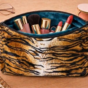 May include: A zippered cosmetic pouch featuring a plush, faux-fur exterior with a brown, black, and white tiger stripe pattern. The interior is lined with a solid teal fabric and contains various makeup items, including brushes and lipsticks.