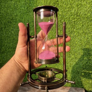 Puede incluir: Un reloj de arena de estilo antiguo con un marco de metal color bronce. El reloj de arena contiene arena rosa y una brújula en la base. El vidrio es transparente y la arena fluye de arriba a abajo. Es un artículo decorativo.