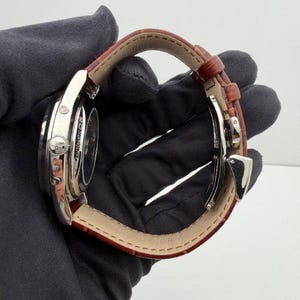 May include: A silver-toned wristwatch with a brown leather band. The watch face is visible, and the band is secured with a silver clasp. The watch is held by a gloved hand.