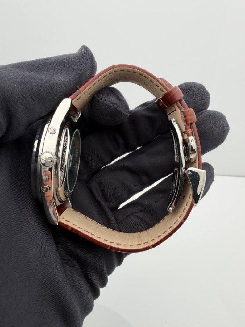 May include: A silver-toned wristwatch with a brown leather band. The watch face is visible, and the band is secured with a silver clasp. The watch is held by a gloved hand.