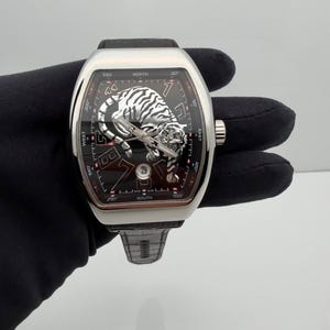 Könnte beinhalten: Eine Luxus-Armbanduhr mit schwarzem Zifferblatt und silbernem Tigerdesign. Die Uhr hat ein rechteckiges Gehäuse mit einem schwarzen Lederarmband. Das Zifferblatt zeigt das Datum und Kompassmarkierungen.