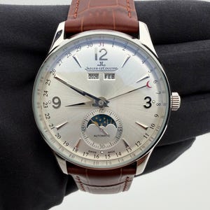 May include: A silver-toned Jaeger-LeCoultre watch with a brown leather band. The watch face displays the date, month, and moon phase. The watch has a silver dial with black numerals and hands.