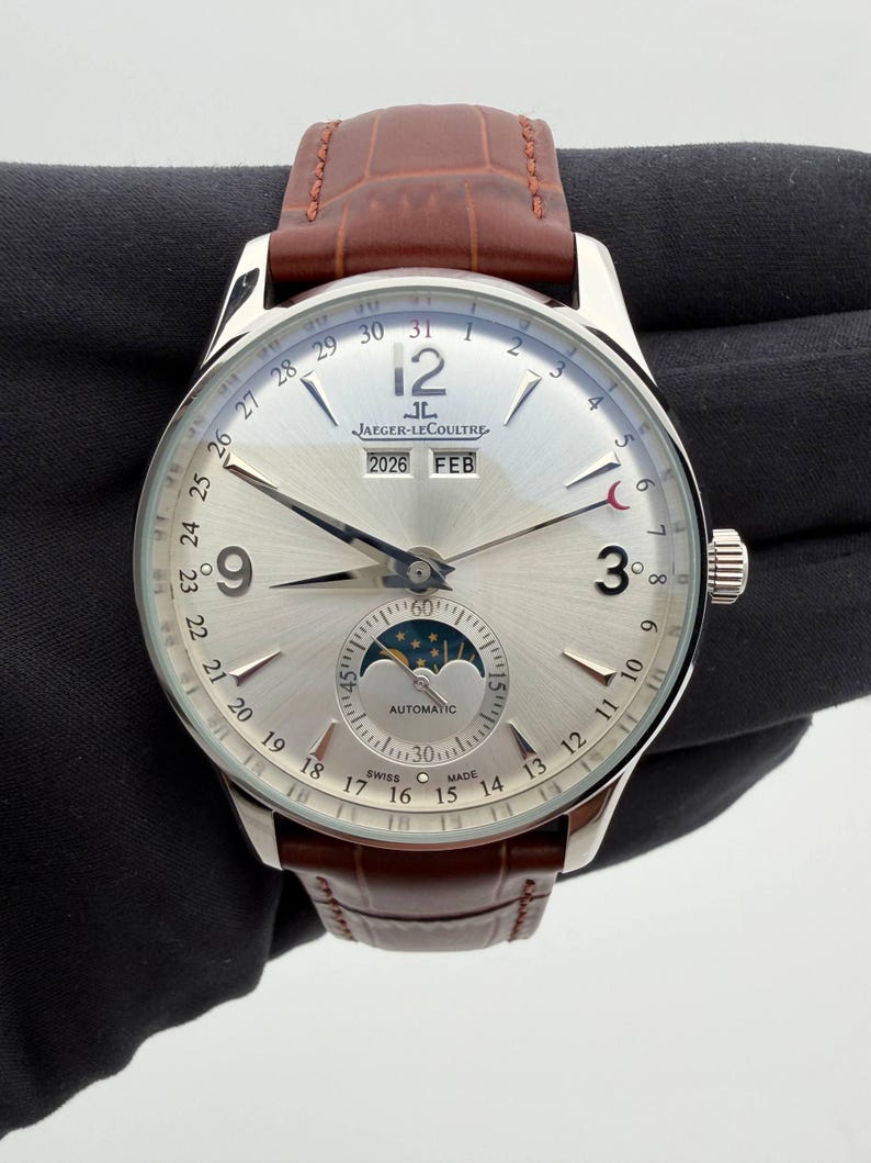 May include: A silver-toned Jaeger-LeCoultre watch with a brown leather band. The watch face displays the date, month, and moon phase. The watch has a silver dial with black numerals and hands.