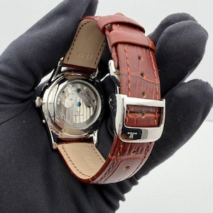May include: A luxury wristwatch with a brown leather strap and a silver buckle. The watch face is visible, showcasing the intricate internal mechanisms. The watch has a silver case and a brown leather strap.