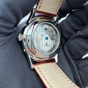 May include: Close-up of a silver-toned wristwatch with a brown leather strap. The watch face is visible through a clear back, showcasing the intricate internal mechanisms. The strap is marked with the size 22-18.