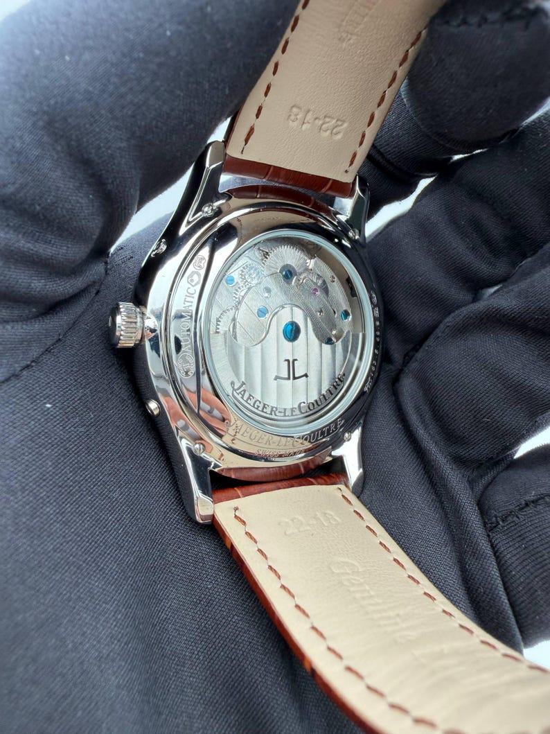 May include: Close-up of a silver-toned wristwatch with a brown leather strap. The watch face is visible through a clear back, showcasing the intricate internal mechanisms. The strap is marked with the size 22-18.