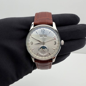 May include: A silver wristwatch with a brown leather strap. The watch face shows the date, month, and moon phase. The watch has the text "JAEGER-LECOULTRE" and "AUTOMATIC" on the face. The watch is held against a dark background.