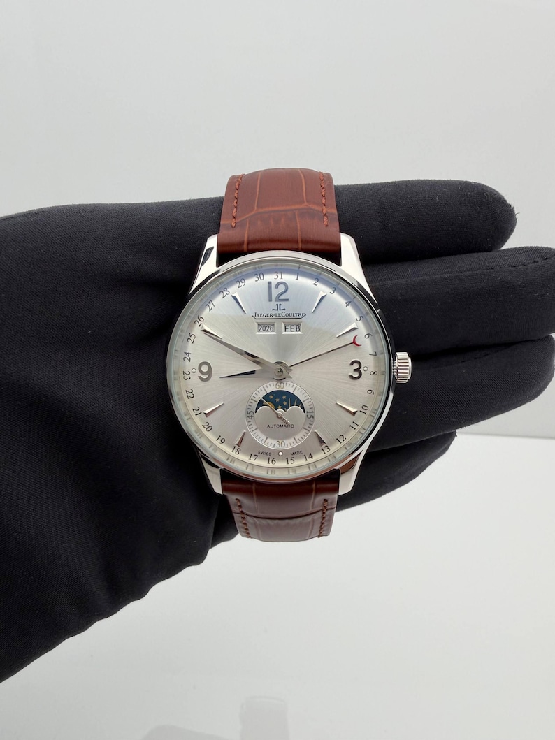 May include: A silver-toned wristwatch with a brown leather band. The watch face displays the date, month, and moon phase. The watch has the text "JAEGER-LECOULTRE" and "AUTOMATIC" on the face. The watch is held against a dark background.