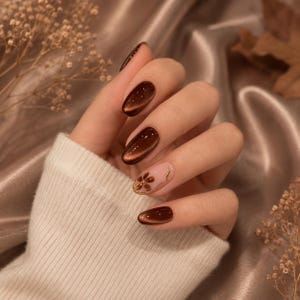 May include: Oval-shaped fingernails featuring a deep, glossy chocolate brown cat-eye polish effect. One accent nail displays a sheer nude base with a small, hand-painted brown flower design and a delicate gold metallic line detail.