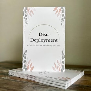 May include: A stack of journals with the title "Dear Deployment" and the subtitle "A Guided Journal for Military Spouses." The cover is white with a semi-circle design and floral accents in gray and peach. The journals are stacked on a wooden surface.