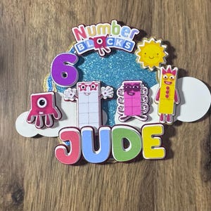 May include: A vibrant Numberblocks-themed birthday decoration. It features the number 6, various Numberblocks characters, a smiling sun, and the name "JUDE". The design incorporates glitter and is set against a wooden background.