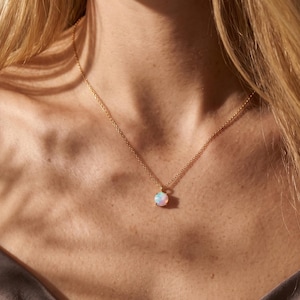 Puede incluir: Un delicado collar de cadena dorada con un colgante de ópalo redondo e iridiscente. La piedra preciosa muestra sutiles destellos de luz azul y rosa, suspendida de una fina cadena de metal en tono dorado pulido que descansa sobre la clavícula.