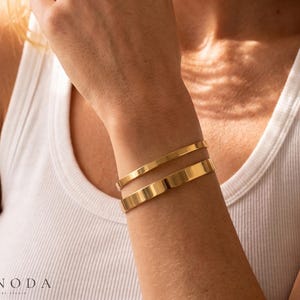 Könnte beinhalten: Zwei polierte goldfarbene Armreifen am Handgelenk. Die Armreifen haben ein glattes, schlichtes Design mit einer reflektierenden metallischen Oberfläche. Der obere Reifen ist schmaler als der untere, beide sind offene, massive Metallbänder.