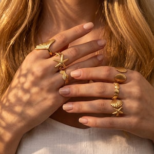 Op de afbeelding: Een collectie goudkleurige ringen met op de oceaan geïnspireerde motieven. De set bevat ringen in de vorm van een waaier, een zeester, een schelp en een band versierd met kleine ronde kraaltjes en een piepkleine zeester.