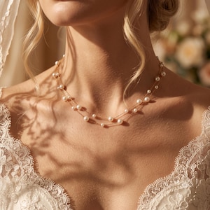 Peut inclure: Un collier délicat à double chaîne dorée orné de perles blanches régulièrement espacées. Le collier repose sur le cou, mettant en valeur une structure à maillons métalliques fins avec de petites perles rondes et lustrées intégrées aux deux chaînes.