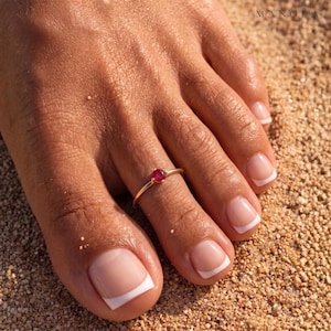 May include: A thin gold toe ring featuring a small, round, faceted red gemstone set in the centre. The ring is worn on the second toe, which has a French manicure with a clear base and a white tip, resting against a textured sandy surface.