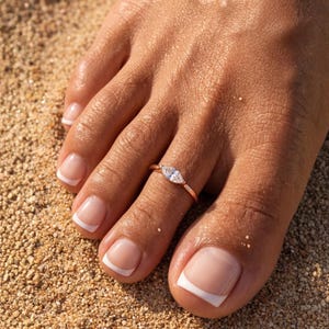 Può includere: Un anello da dito del piede in oro rosa con una singola pietra preziosa trasparente a taglio marquise, incastonata orizzontalmente su una sottile fascia metallica lucida. L'anello è indossato sul secondo dito, poggiato su sabbia granulosa.