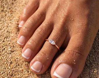 May include: A rose gold toe ring featuring a single marquise-cut clear gemstone set horizontally on a thin, polished metal band. The ring is worn on the second toe, which is positioned against a textured, sandy surface with visible grains.