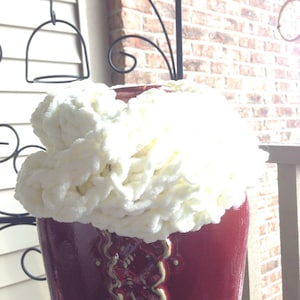 May include: A white crocheted scarf sits on top of a red ceramic vase with a gold and white pattern. The vase has a rounded shape and a wide opening.