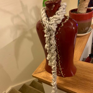 Puede incluir: Una bufanda de crochet blanca drapeada sobre un jarrón de cerámica rojo con detalles dorados. El jarrón está sobre una superficie de madera.